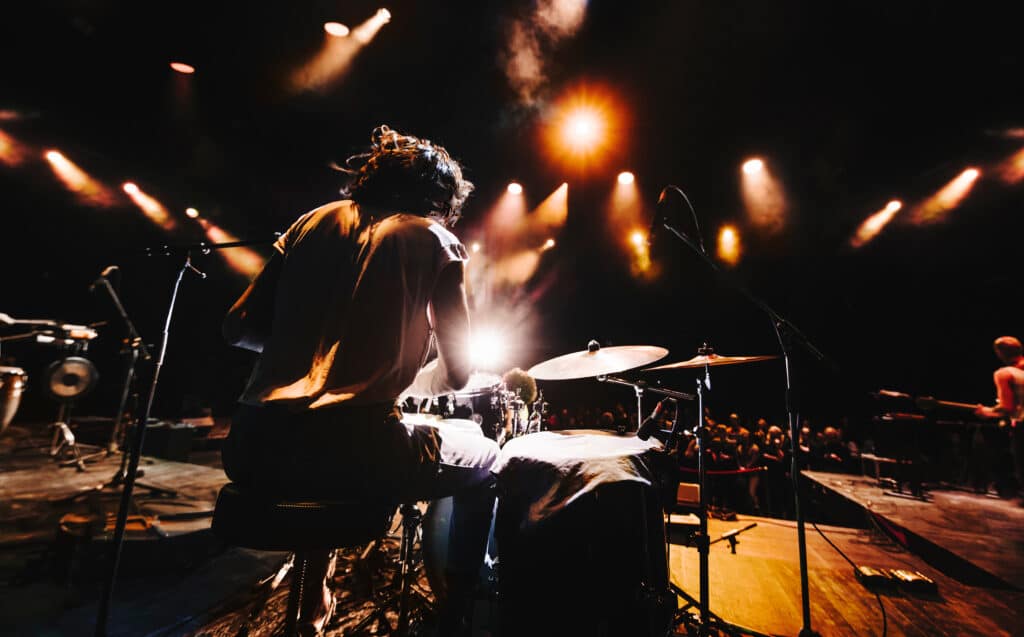Batteur sur scène vu de dos, sous les projecteurs et la fumée, ambiance de concert live intense avec foule.