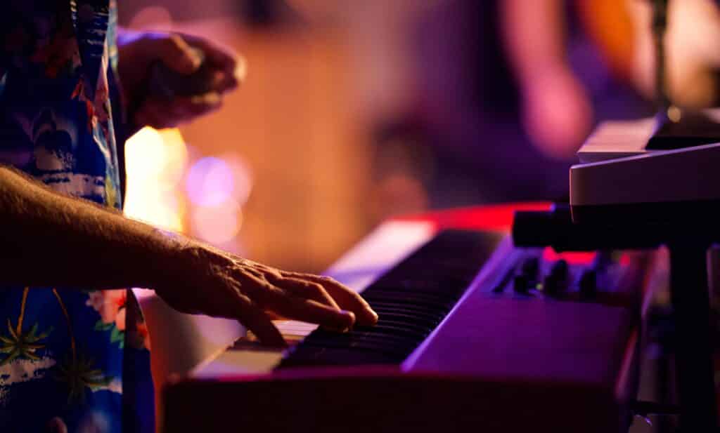 Mains jouant sur un clavier électronique, ambiance de concert aux lumières violettes et orangées. Chemise à motifs tropicaux.