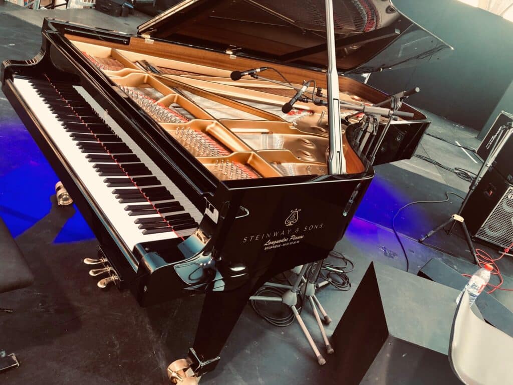 Piano à queue Steinway & Sons noir brillant sur scène, capot ouvert. Microphones sur les cordes. Texte visible : Languedoc Pianos 04 67 35 36 88.