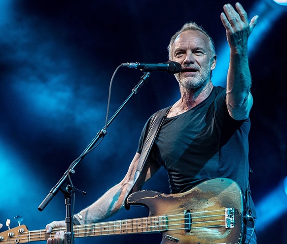 Sting sur scène joue de la basse et chante dans un micro, bras levé, lumière bleue.